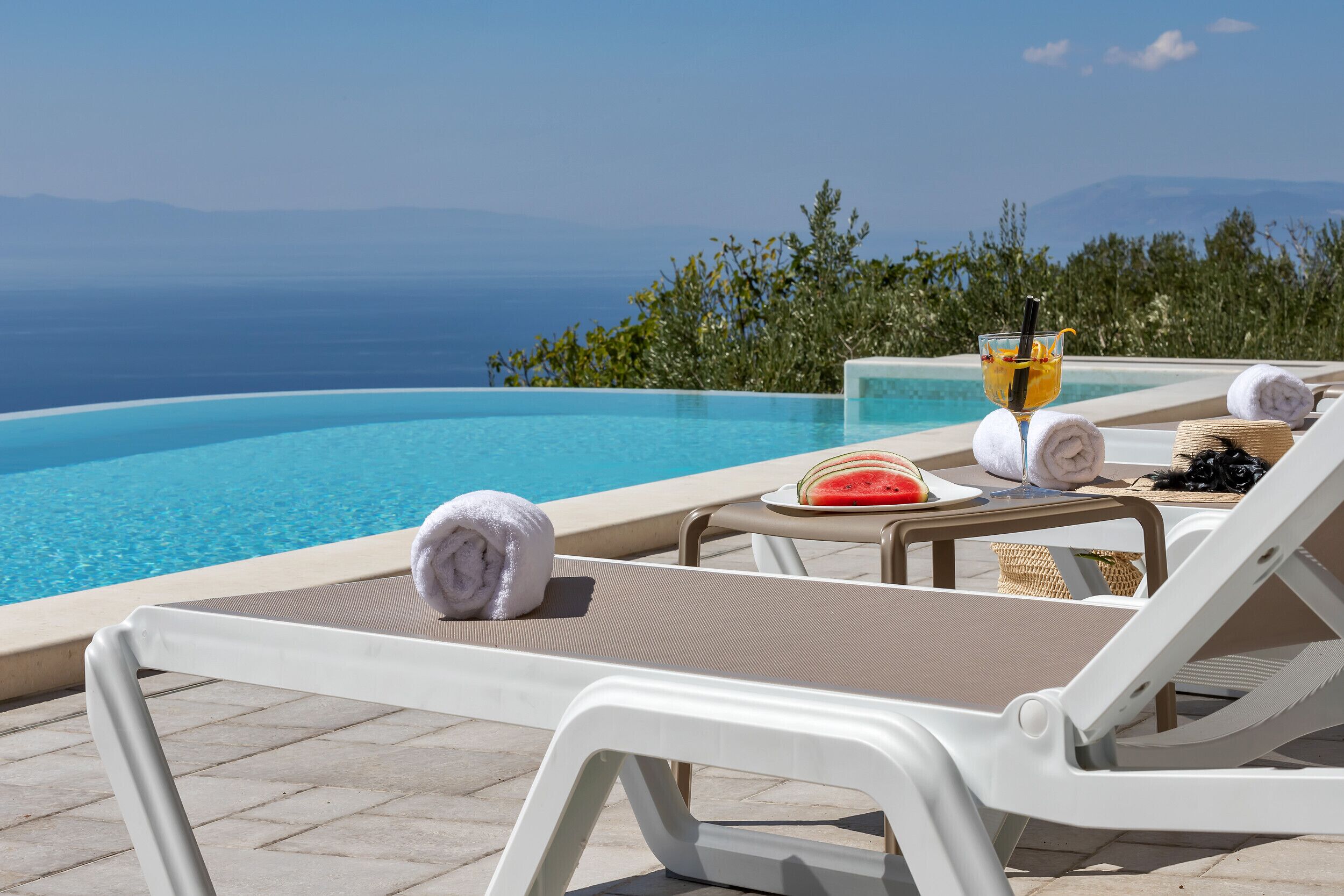 A poolside lounge with a view of the ocean, a glass of drink, and a watermelon on a table.