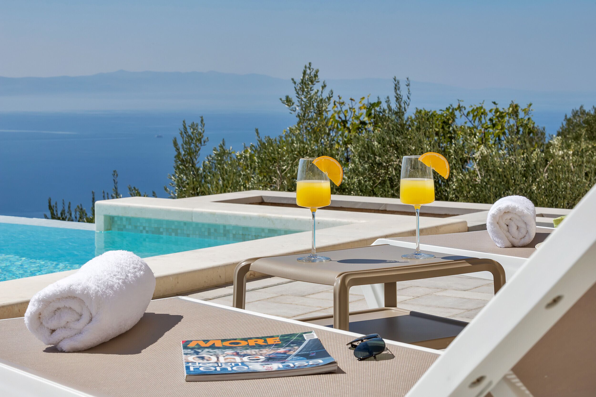 A poolside lounge with two glasses of orange juice, a rolled towel, and a magazine on a table.