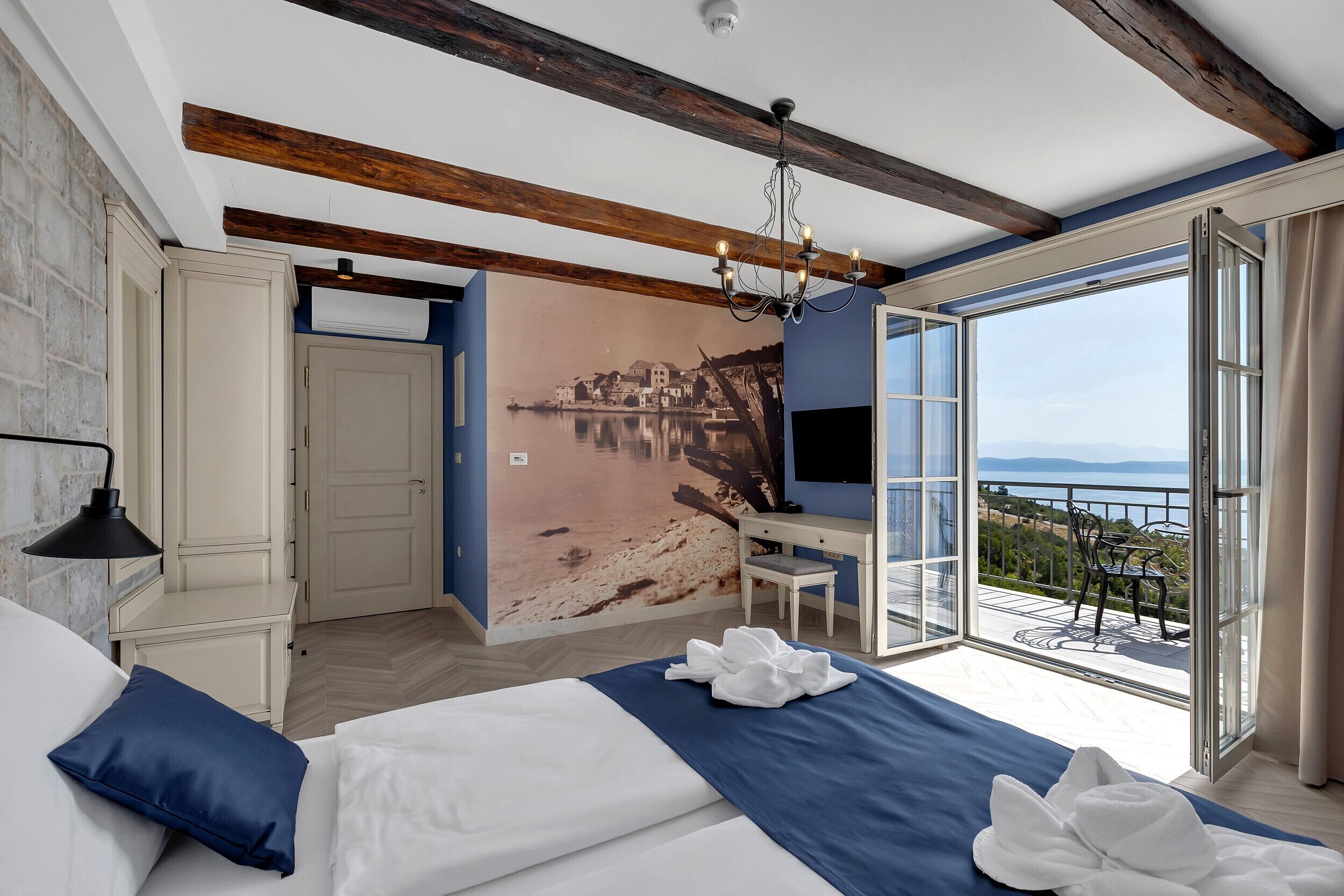 A bedroom with a large bed, a desk, and a view of a balcony with outdoor seating.