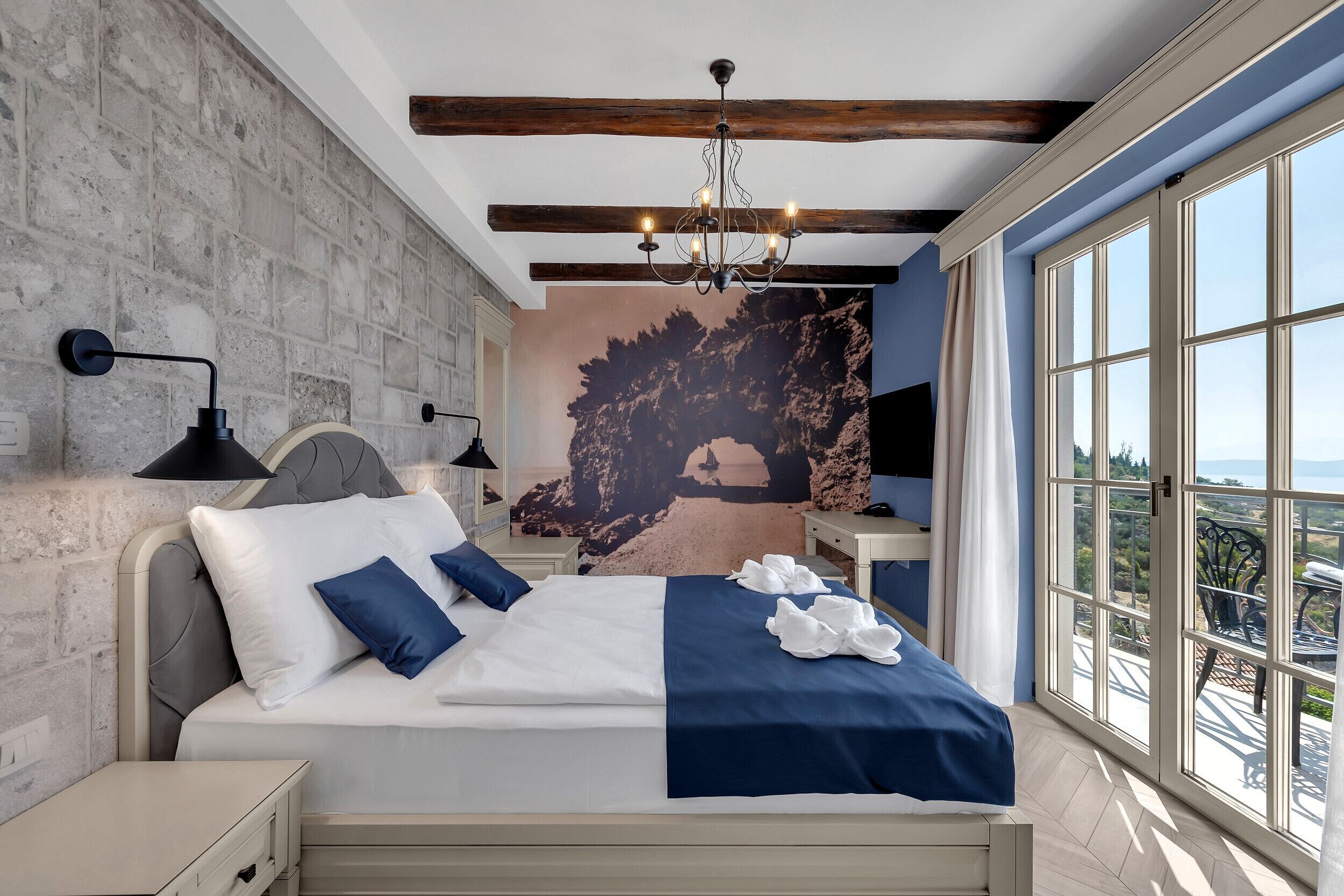 A bedroom with a bed, a nightstand, a wall-mounted lamp, a large window with a view, and a chandelier.