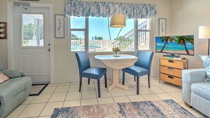 A living room with a dining table, two chairs, a TV, and a view of the beach.
