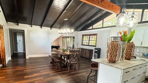 A spacious loft with a high ceiling, dark wooden beams, a dining area with a wooden table and chairs, and a kitchen with white cabinets and a large window. - Unique Spanish Style Lakefront Home!  Kayaks and Paddle Boat! (Keystone Heights)