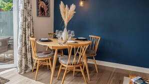 A dining area with a round wooden table set for four, surrounded by wooden chairs with patterned cushions. A vase with pampas grass and a wall-mounted light fixture are visible. - Cute & Cosy Cottage | Chilterns - Pass the Keys (Princes Risborough)