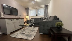 Un salon moderne avec un canapé gris, une table basse en verre, un meuble blanc, un téléviseur à écran plat fixé au mur et une fenêtre avec des rideaux.