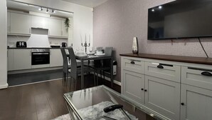 Une cuisine moderne avec des meubles blancs, un coin repas doté d’une table en verre et un téléviseur à écran plat fixé au mur.
