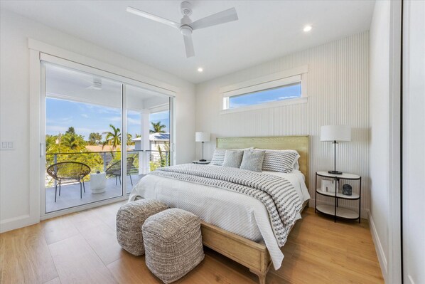 A bedroom with a large bed, a ceiling fan, a window, and a balcony with outdoor furniture.