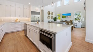 A modern kitchen with white cabinetry, a large island, stainless steel appliances, pendant lighting, and a view of the living area with a sofa and a painting. - AMI Escape! Swim-Up Bar, Ping Pong, Spa and Walk to Beach (Anna Maria)