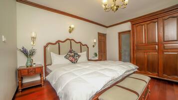 A bedroom with a large bed, wooden wardrobe, and a nightstand with a vase and lamp.
