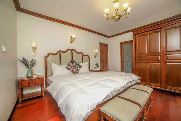 A bedroom with a large bed, wooden wardrobe, and a nightstand with a vase and lamp.