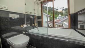 A bathroom with a bathtub, toilet, and a window with a view of houses. - The Winterfell (Kotagiri)