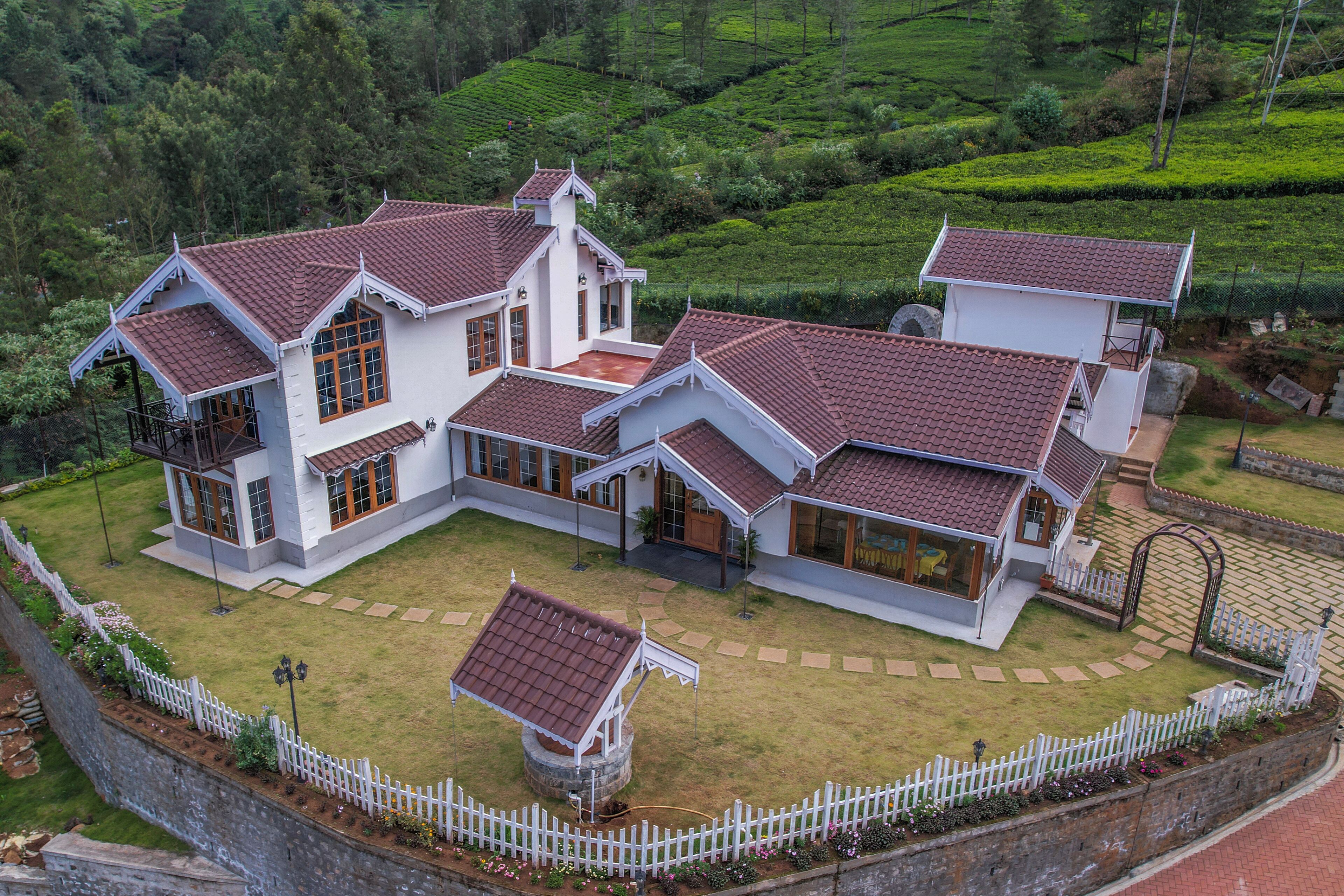 A large, white, two-story house with a red-tiled roof, surrounded by a white picket fence and lush green tea plantation in the background.