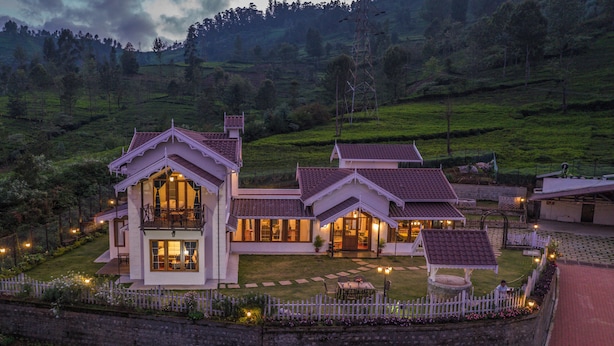 A large, multi-story house with a white exterior and red-tiled roof, surrounded by a white picket fence and lush greenery. - The Winterfell (Kotagiri)