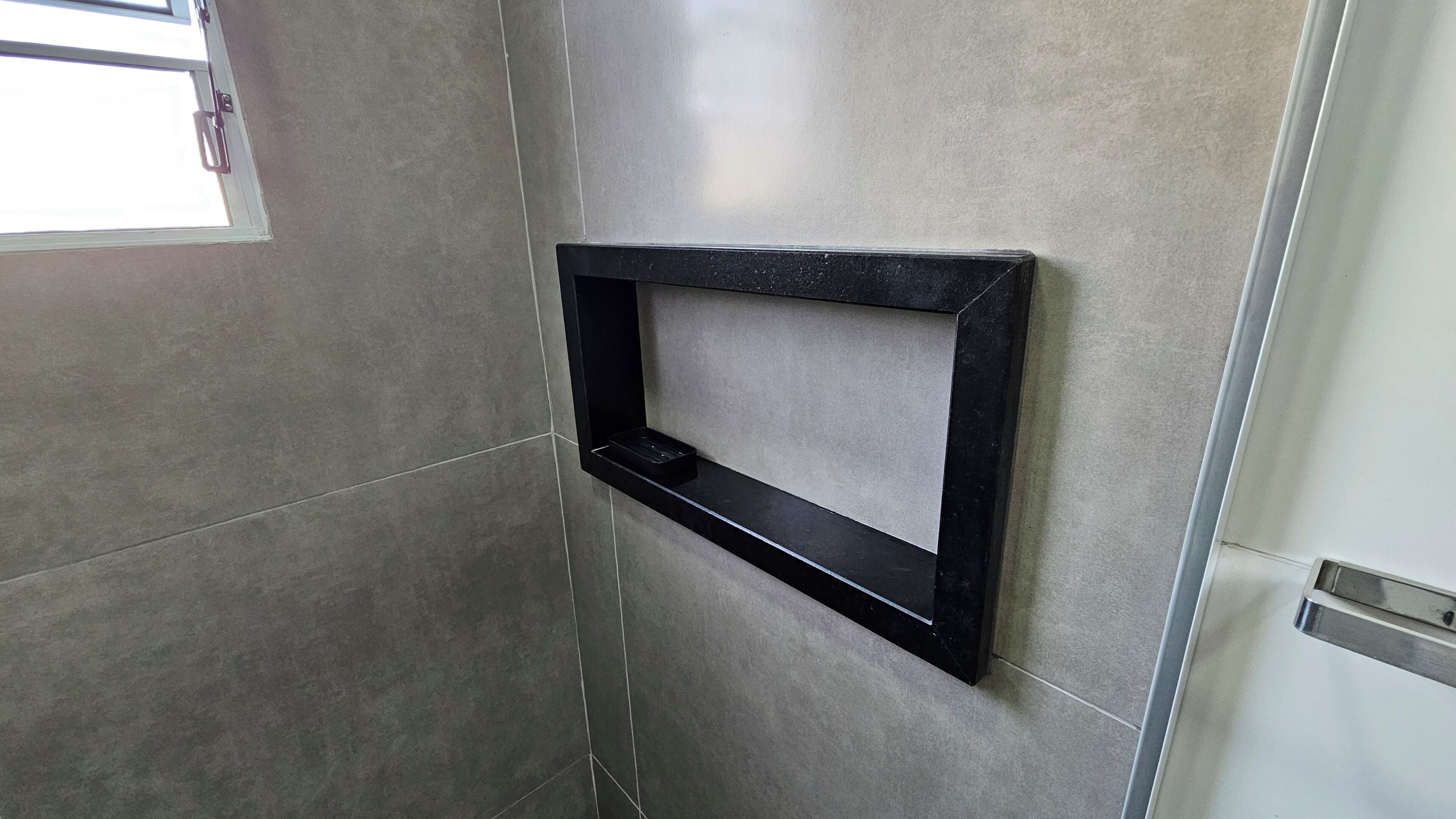 Une salle de bain avec des murs recouverts de carreaux gris, un miroir à cadre noir et une porte dotée d’une poignée métallique.