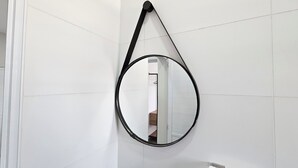 Un baño con paredes de azulejos blancos, un espejo redondo con marco negro y un banco de madera.