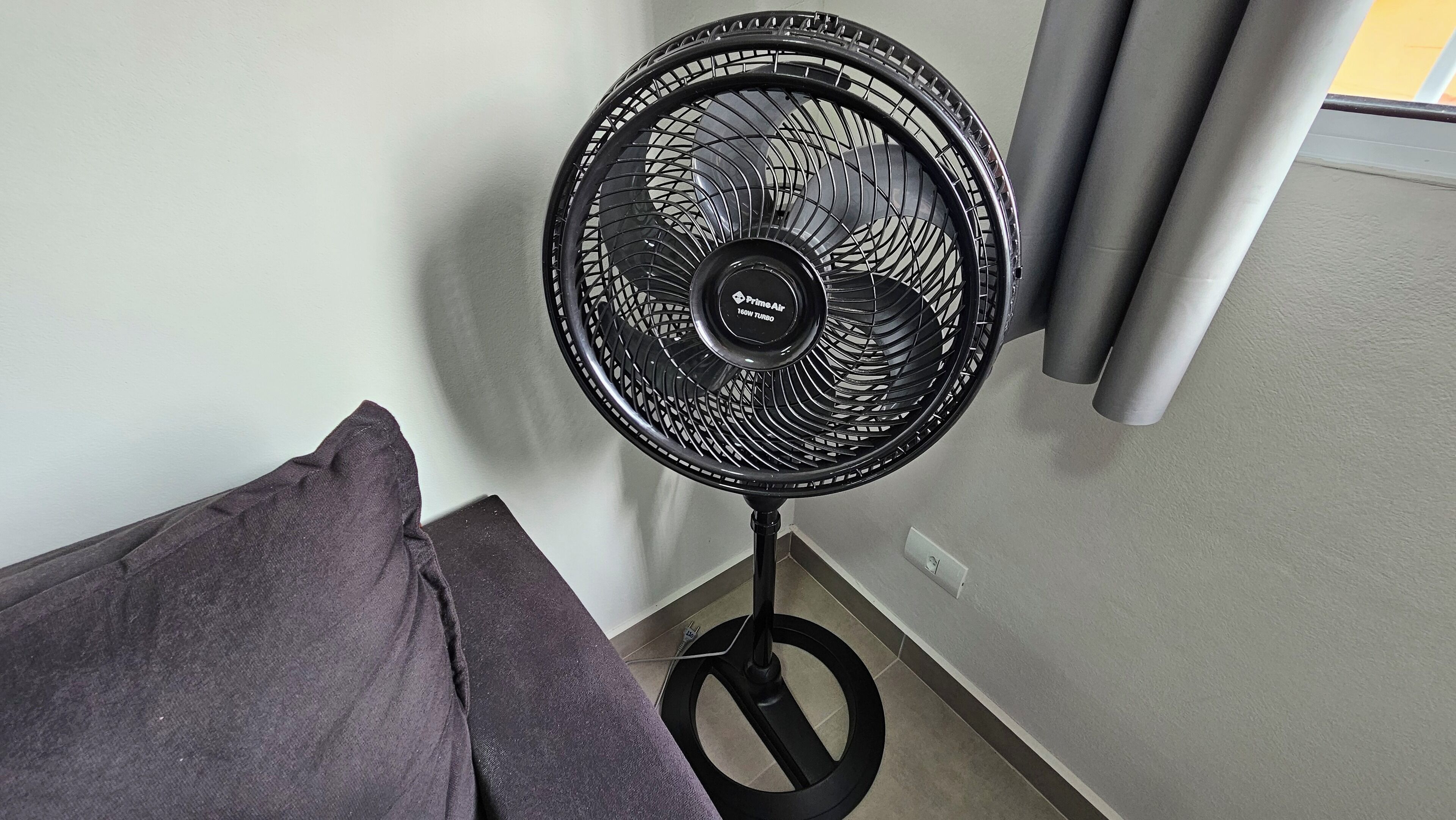 A standing fan with a circular design, positioned against a wall with a light switch nearby.