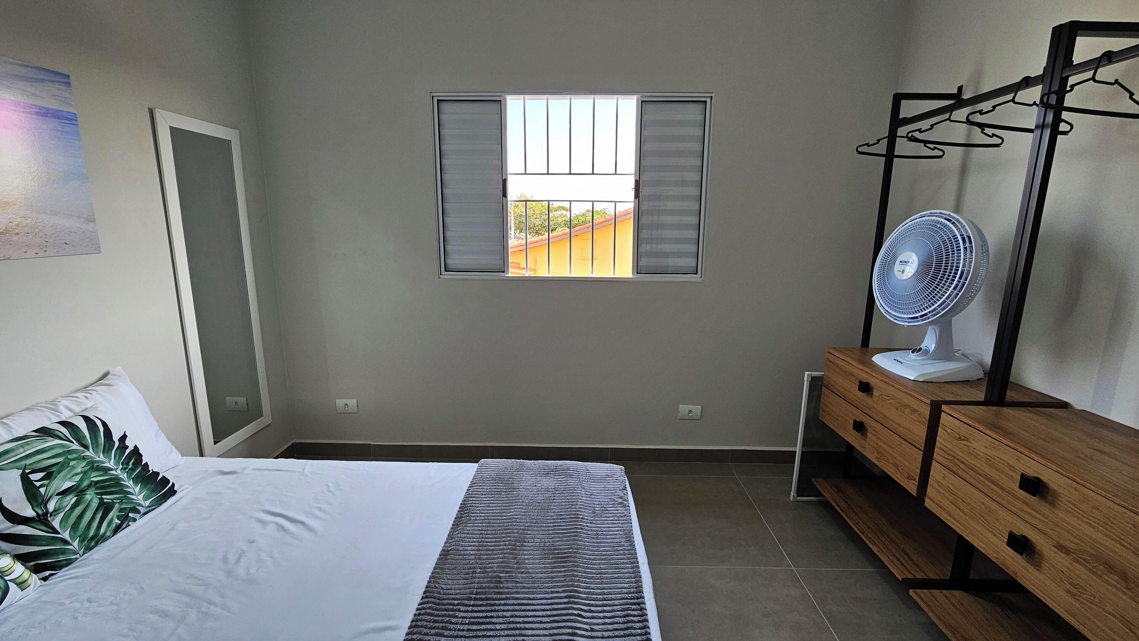 Un dormitorio con cama, mesita de noche con ventilador, ventana con persianas y vista a un edificio.