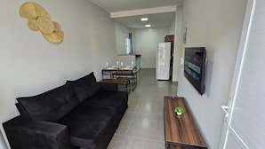 Una sala de estar moderna con un sofá negro, una mesita de madera con una planta, y una cocina al fondo.