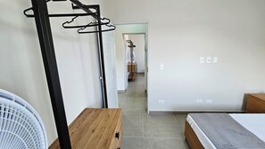 A bedroom with a bed, a wooden nightstand, a wall-mounted fan, and a hallway leading to a bathroom with a towel rack. - Spazio Italia Apartamentos (Ilha Comprida)