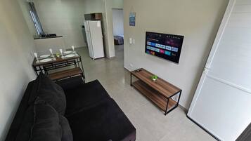 Un espacio habitable pequeño con un sofá, una pequeña mesa de comedor, un televisor montado en la pared y un refrigerador al fondo.