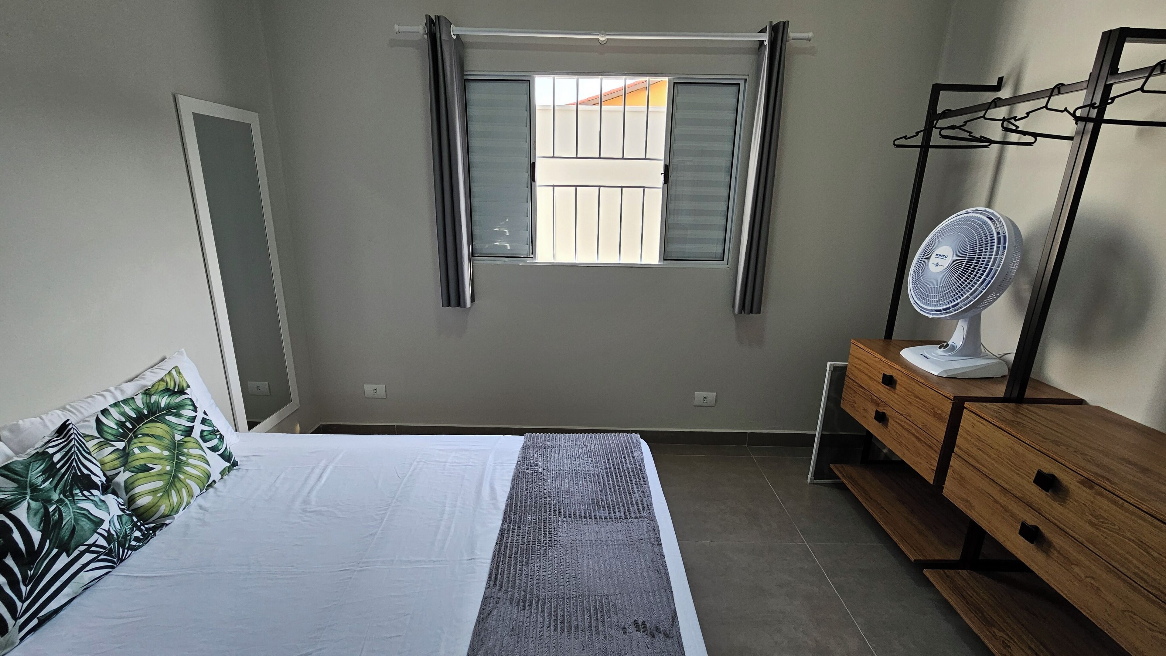 Une chambre à coucher comprenant un lit, une commode, un miroir, une fenêtre avec des stores et un ventilateur sur pied.