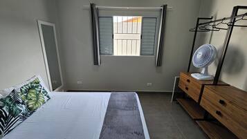 Une chambre à coucher comprenant un lit, une commode, un miroir, une fenêtre avec des stores et un ventilateur sur pied.