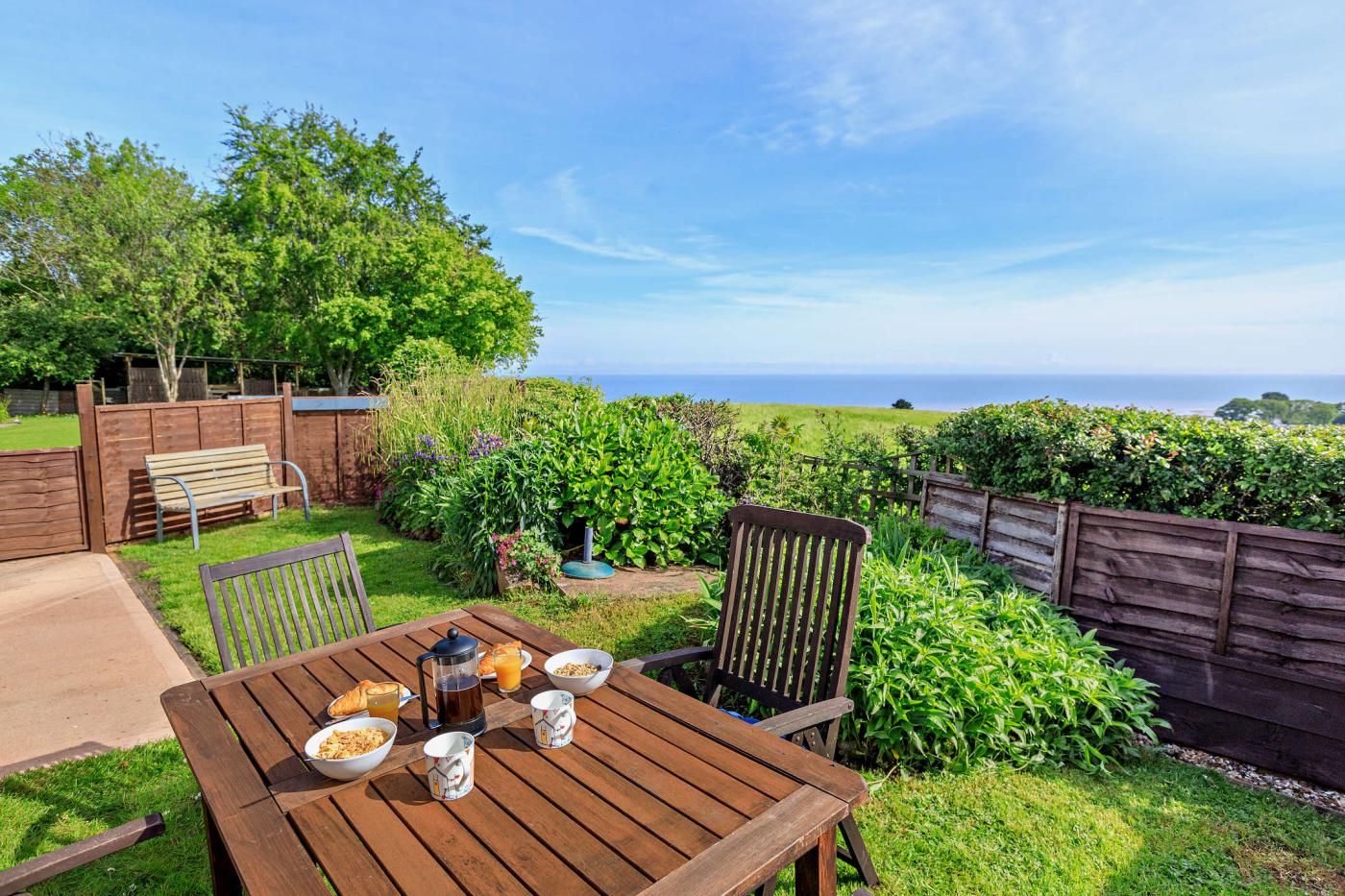 Ein Holztisch ist reichhaltig mit Frühstücksleckereien gedeckt, umgeben von Stühlen, in einem Garten mit Blick auf das Meer.