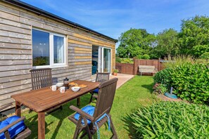 A wooden cabin with a dining table, chairs, and a garden area with a bench and potted plants.
