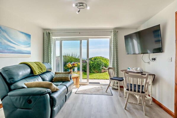 A living room with a leather sofa, a dining table with chairs, a flat-screen TV, and a sliding glass door leading to a garden.