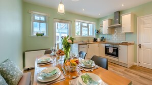 A dining area with a wooden table set for a meal, a kitchen with white cabinets, and a view of the outside through the windows. - 3 Bed in Braunton (oc-b29581) (Braunton)