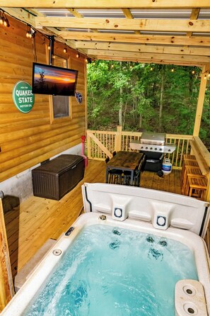 Una terraza de madera con un jacuzzi, una parrilla y una pantalla de televisión.