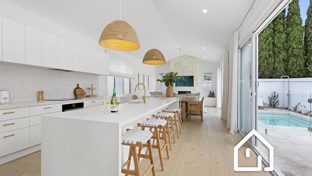 Une cuisine moderne avec des meubles blancs, un îlot central doté de tabourets de bar, un éclairage suspendu et une vue sur une piscine à l’extérieur.
