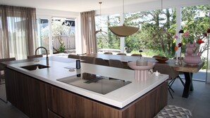 A modern kitchen with a large island, a dining area with chairs, and a view of trees through large windows.
