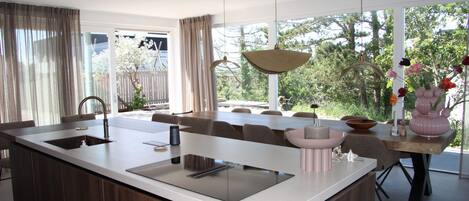 Een moderne keuken met een groot eiland, een eethoek met stoelen, en uitzicht op bomen door grote ramen.
