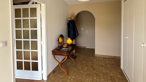 Un couloir doté d’un sol carrelé en forme d’hexagone, une console en bois avec un miroir, une lampe et un panier. On y trouve une porte avec un panneau de verre, un porte-manteau avec une veste, et une armoire blanche.