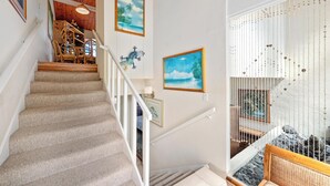 Una escalera con barandilla de madera, vista de una habitación con cama y ventana, y una pared decorada con obras enmarcadas.