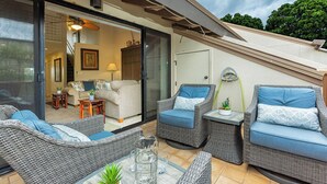 An outdoor patio with wicker furniture, a glass door leading to an interior living area, and a view of trees. - Maui Kamaole M207 Kihei condo w/ pool, hot tub and sunset lanai, summer deal (Kihei)