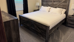 Une chambre à coucher avec un lit en bois, une table de chevet avec une lampe, une fenêtre avec des rideaux noirs et un luminaire fixé au mur.