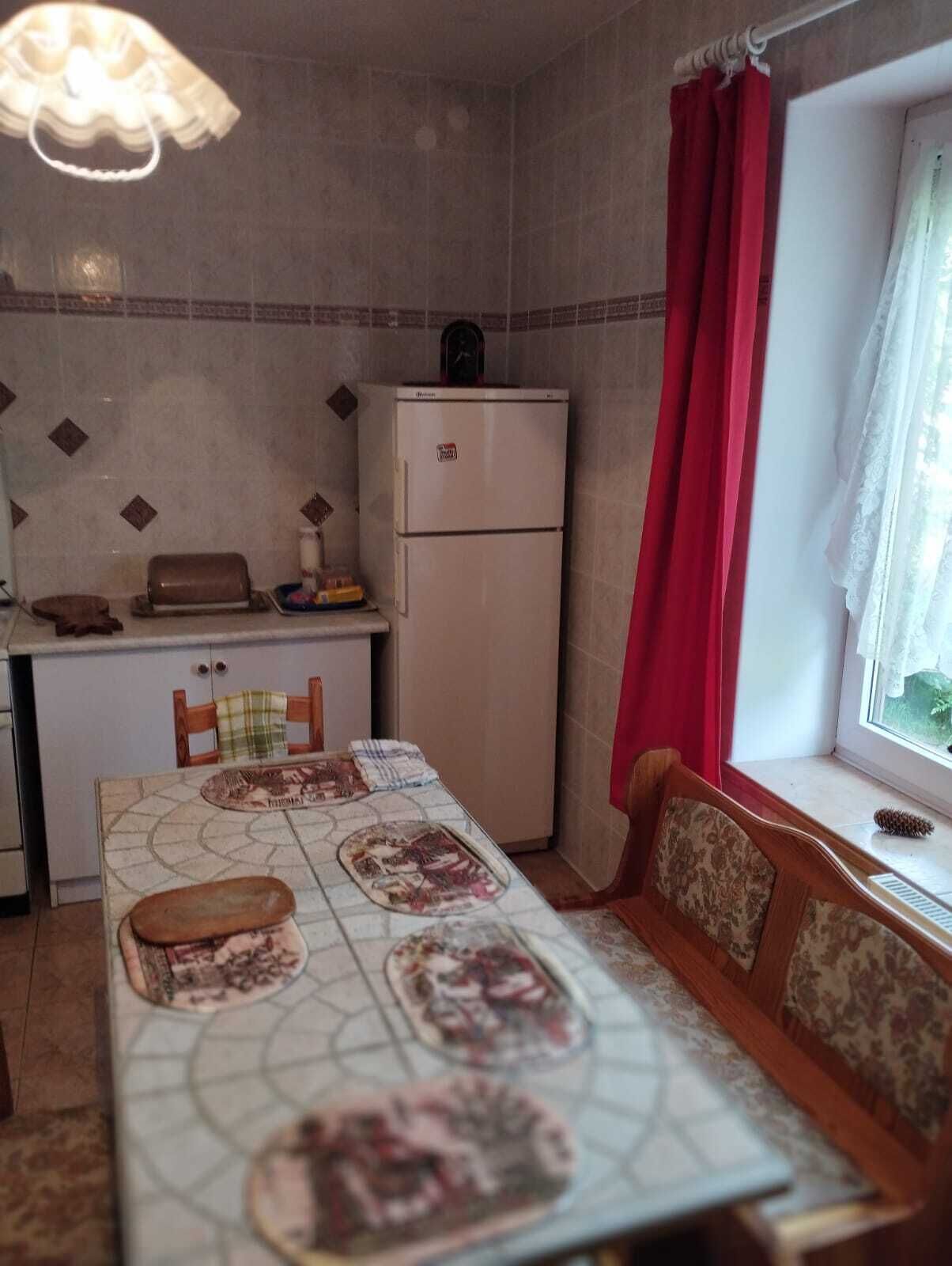 A compact kitchen with a refrigerator, a dining table with a patterned tablecloth, and a window with red curtains.
