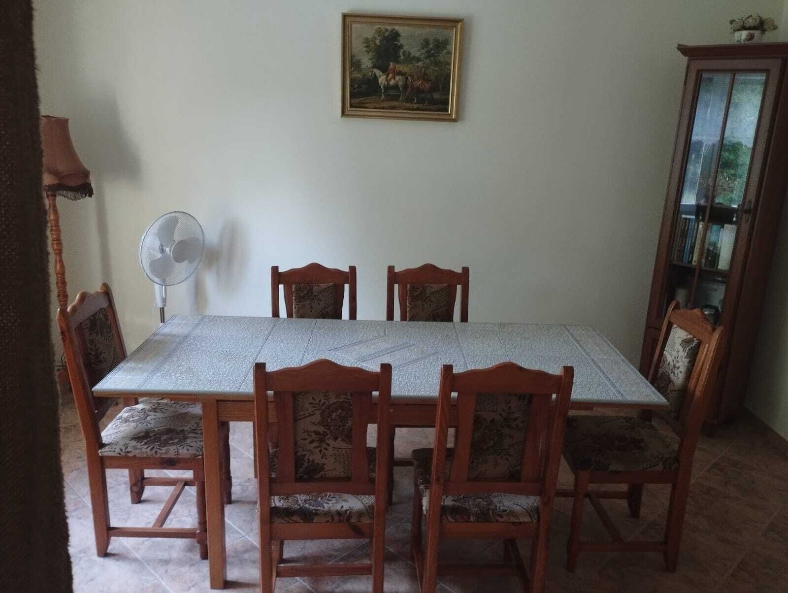 Un área de comedor con una mesa rectangular, seis sillas de madera con cojines estampados, un ventilador de pared y un cuadro enmarcado sobre la mesa.