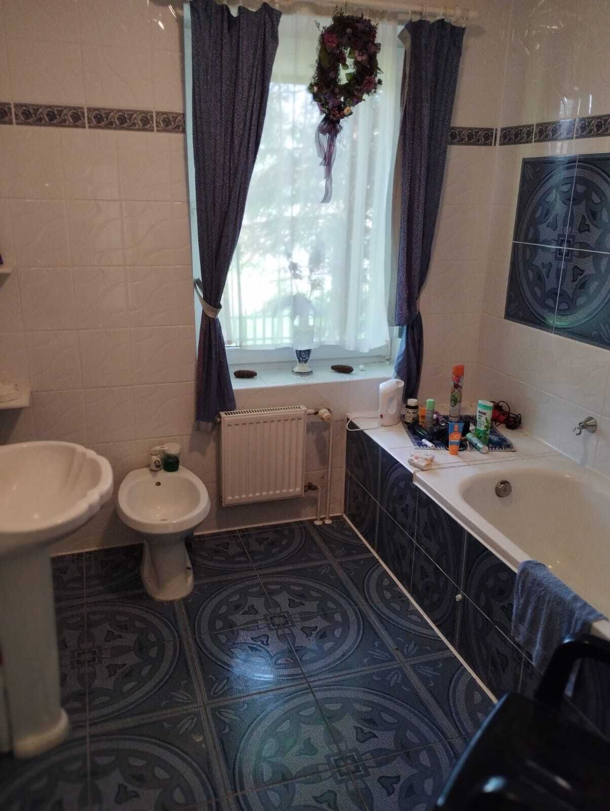 Ein Badezimmer mit einer Badewanne, einer Toilette und einem Bidet. Ein Fenster mit Vorhängen und ein Kranz daran.