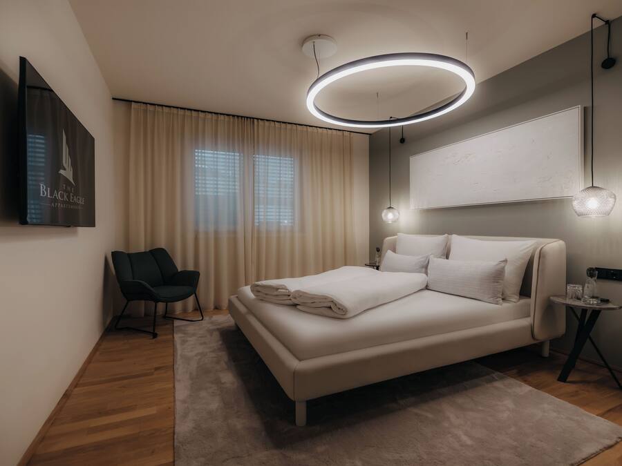 A modern bedroom with a bed, a chair, a bedside table, and a wall-mounted TV.
