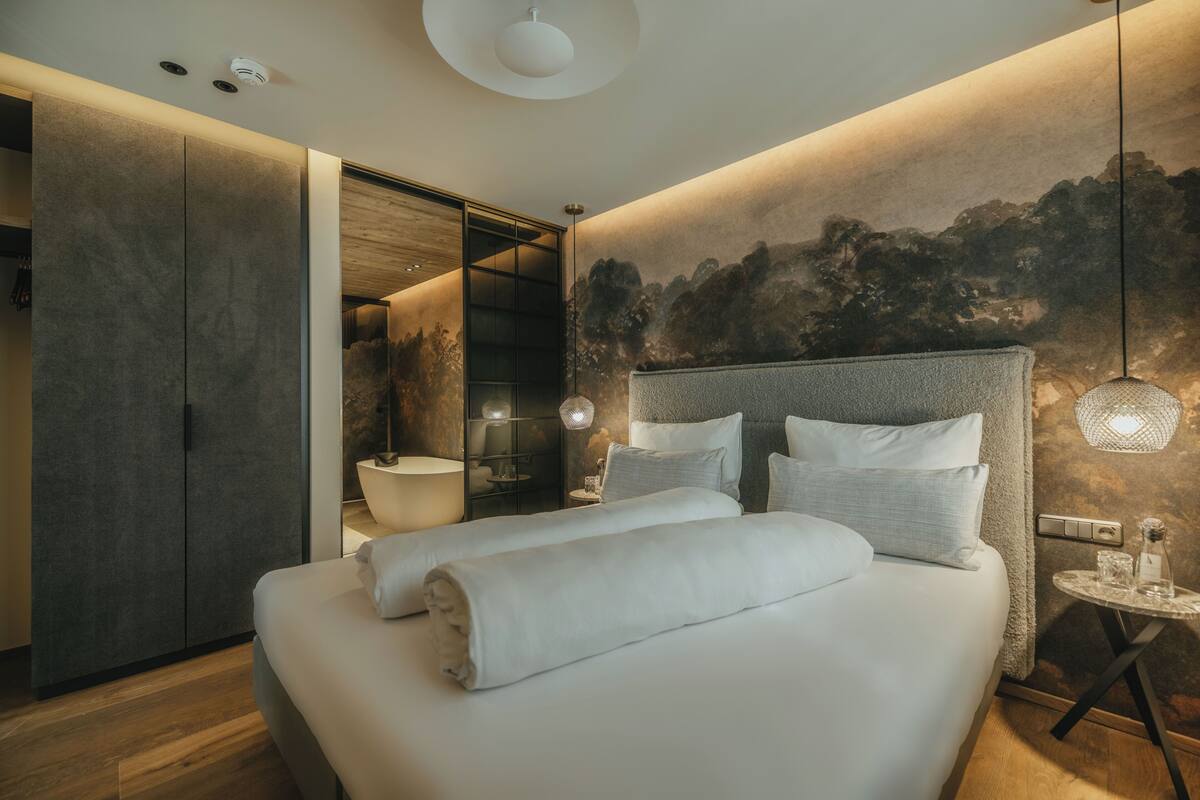 Un dormitorio moderno con una cama grande, piso de madera, cabecera fijada a la pared y una pequeña mesa redonda con lámpara.
