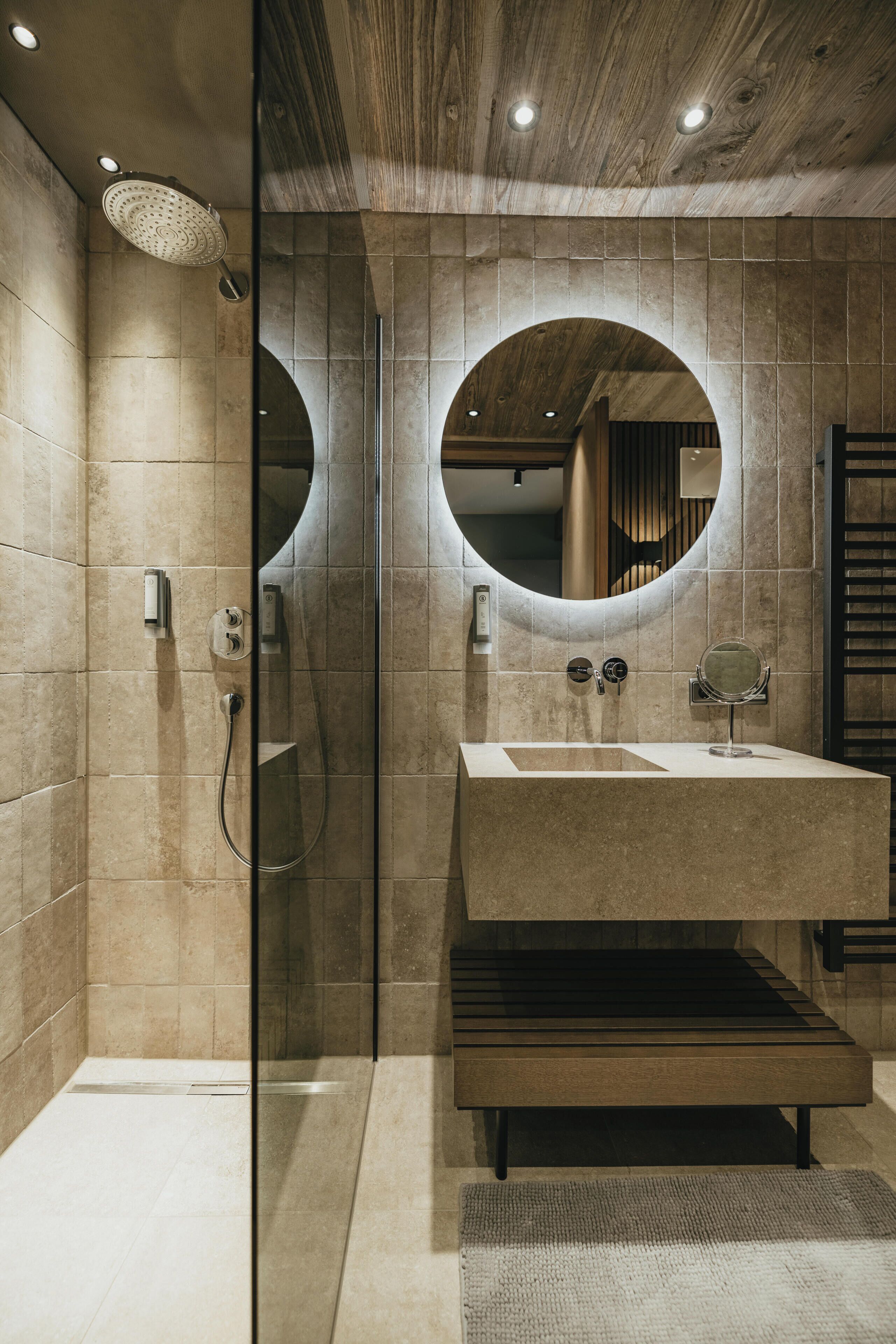 Ein modernes Badezimmer mit einer Glaskabine zur Dusche, einem großen Waschbecken mit rundem Spiegel und einer hölzernen Sitzbank.