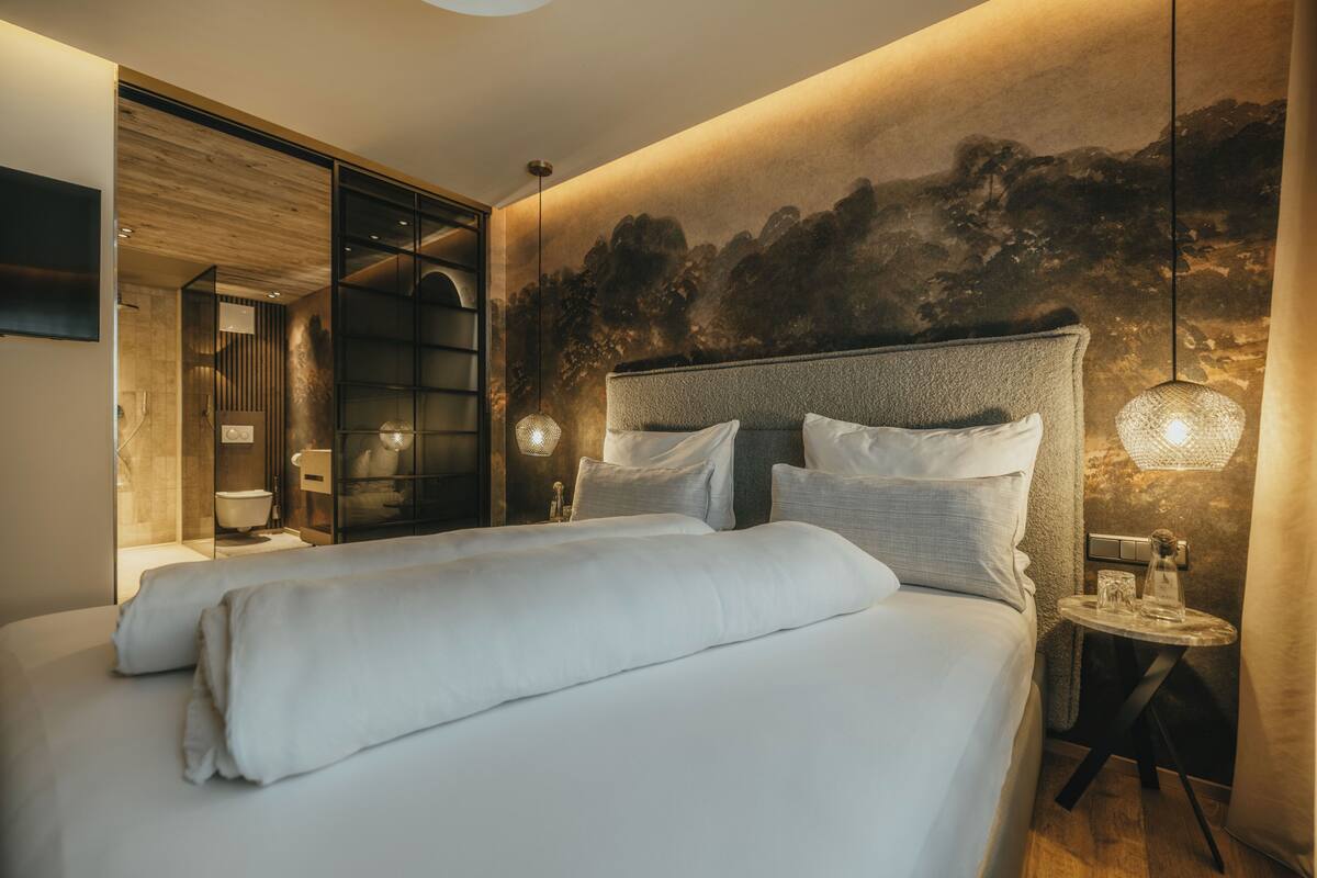 Un dormitorio moderno con una cama grande, una mesita de noche, una lámpara de pared y un cuadro de un bosque en la pared.