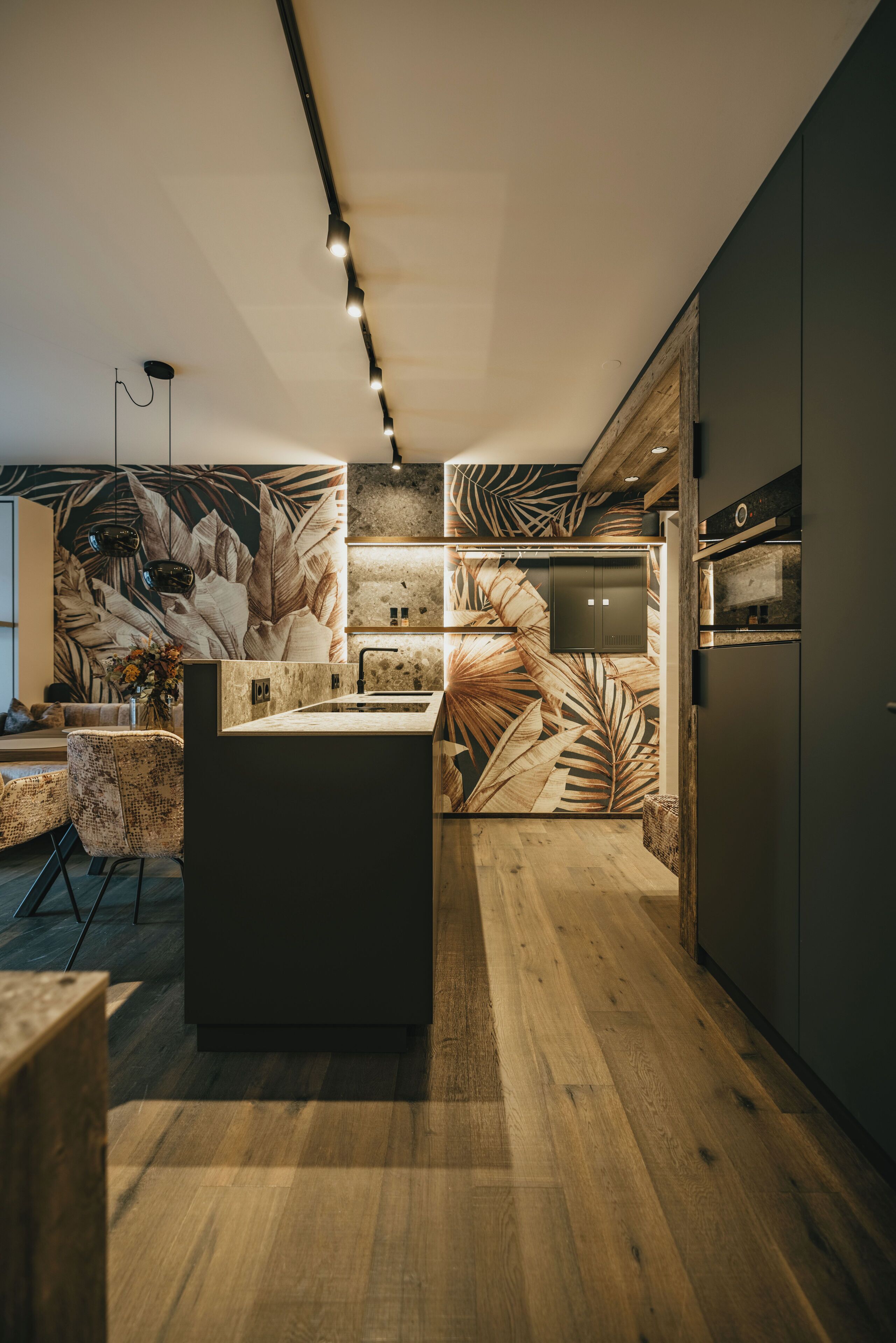 A modern kitchen with a central island, wooden flooring, and a tropical leaf-patterned backsplash.