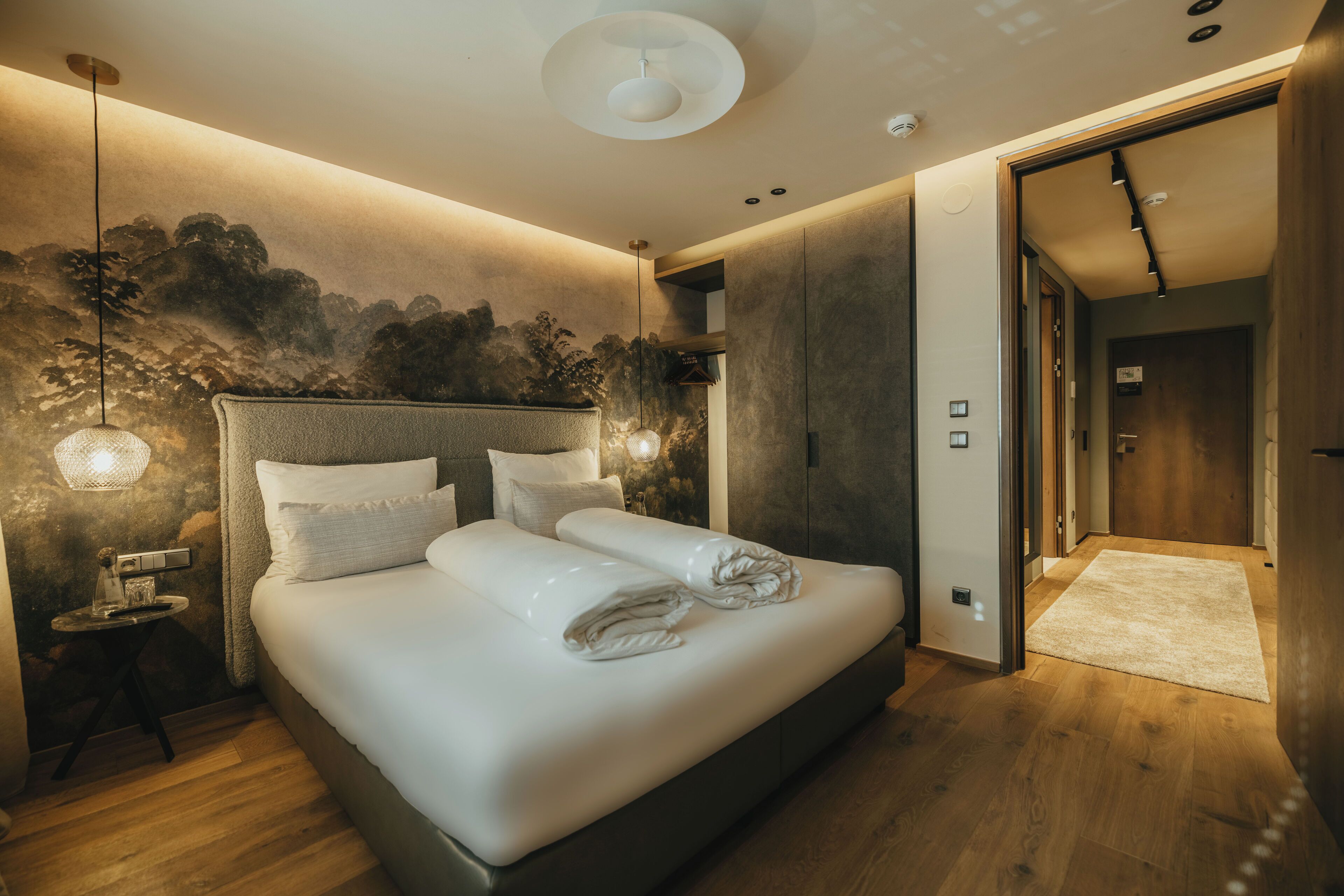 Un dormitorio moderno con una cama grande, una mesita de noche, una lámpara de pared y una puerta que da a otra habitación.