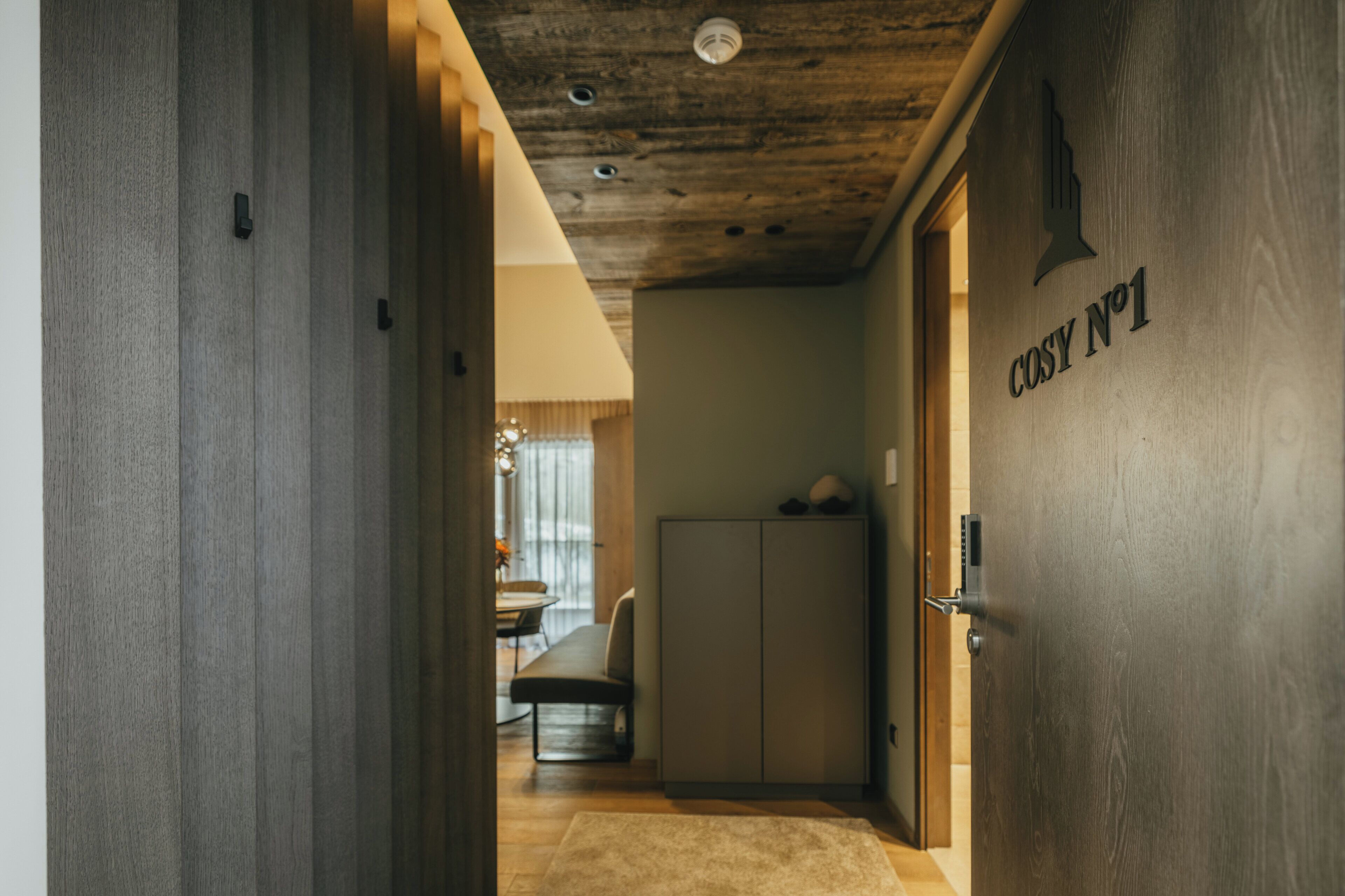 A hallway with a wooden door labeled "COSY No 1", a kitchen area, and a dining space with a table and chairs.