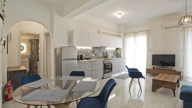 A modern living room with a glass dining table, blue chairs, a wooden coffee table, a TV, and a kitchen area with white cabinets and a tiled backsplash.