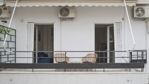 A balcony with two wicker chairs and a table, overlooking a building with two open doors and air conditioning units.