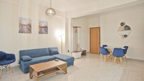 A modern living room with a blue sofa, a wooden coffee table, and a dining area with blue chairs. There are abstract paintings on the walls and a mirror with decorative elements.